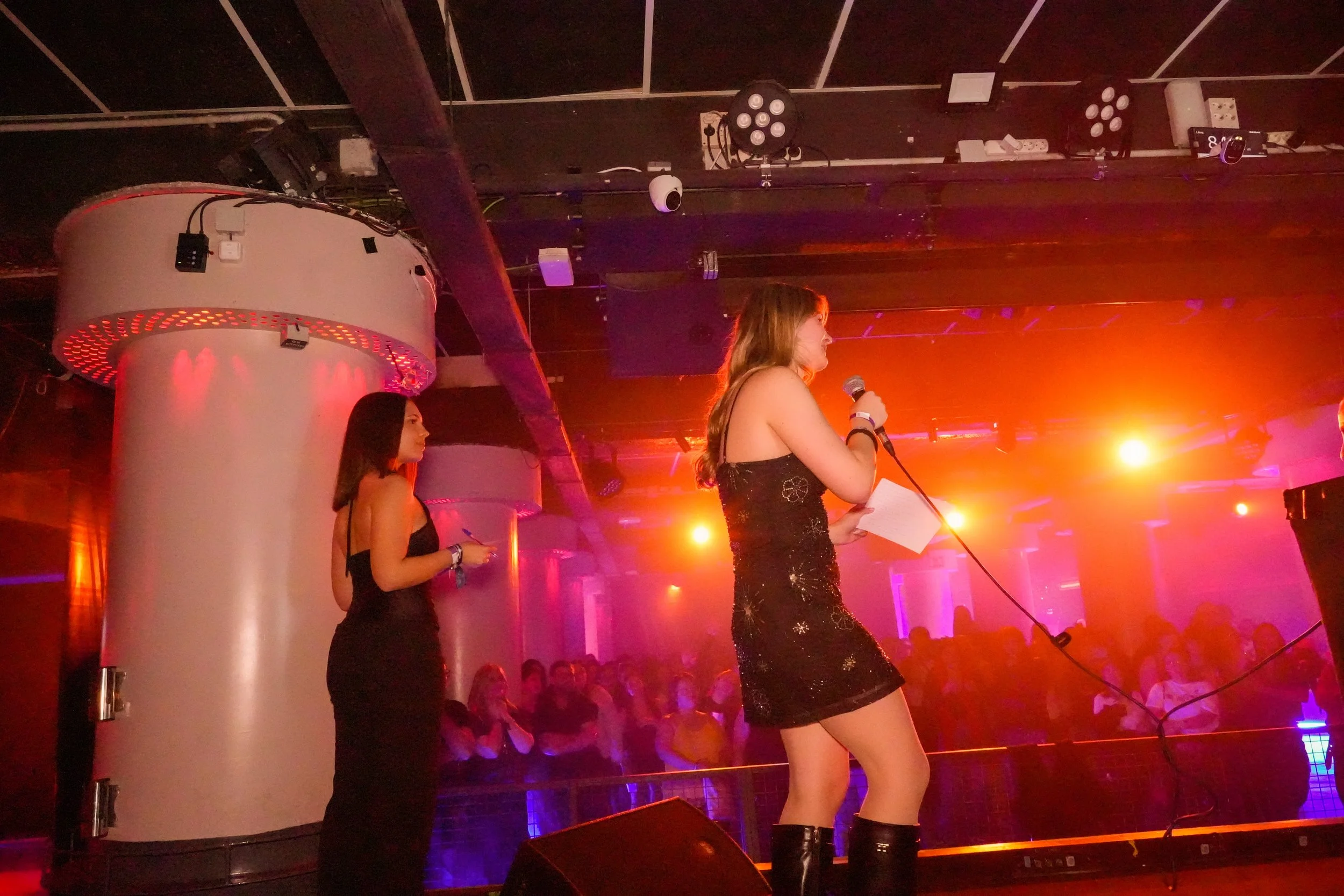 Two women standing on stage in a dimly lit venue with red and purple lighting. One woman is speaking into a microphone, while the other looks on. Audience members are visible in the background.