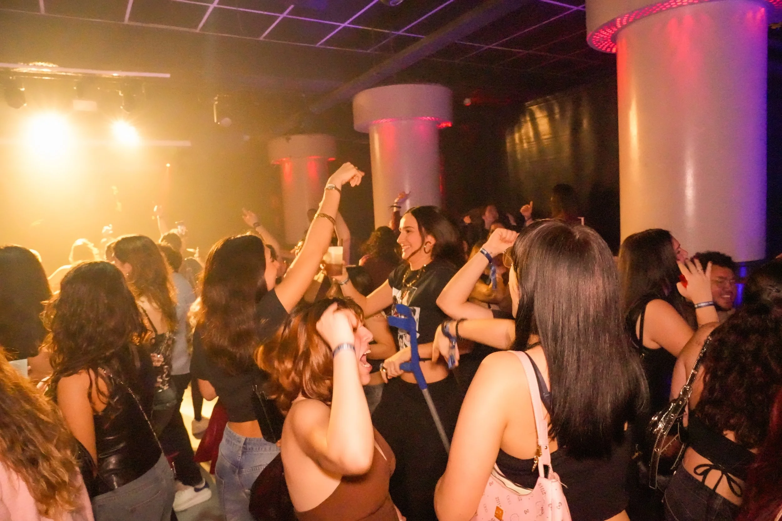 People dancing and enjoying music at a nightclub with colorful lighting and large pillars.