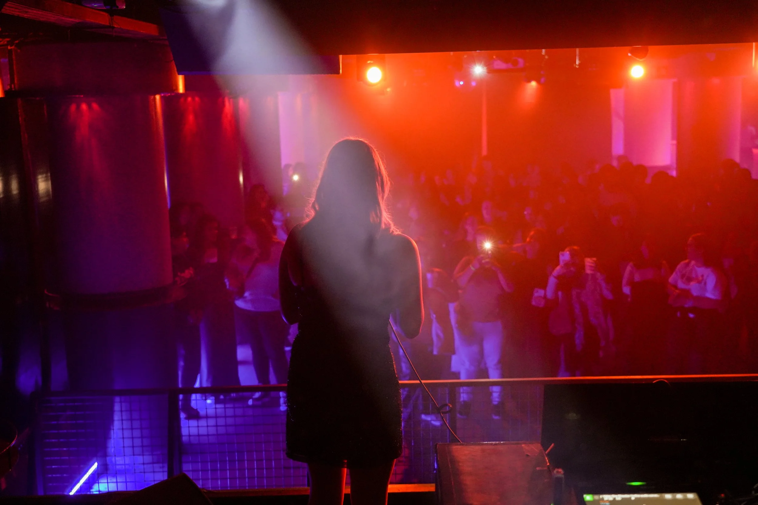 A woman with long hair standing on a stage in front of a large audience at a nightclub or concert venue, illuminated by colorful stage lights.