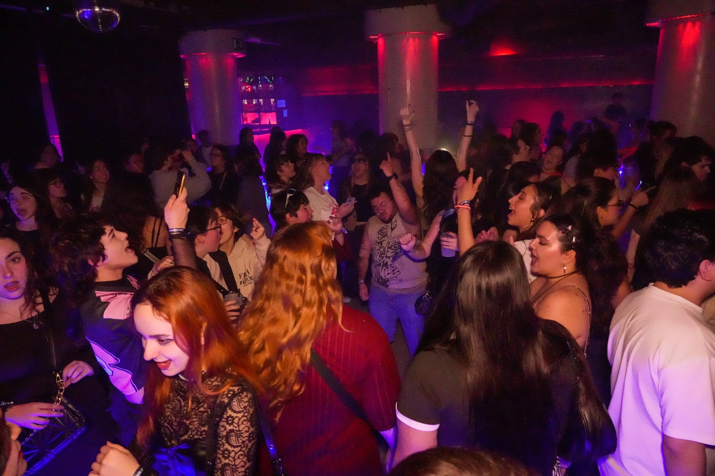 Crowded dance floor at a nightclub with people dancing, socializing, and enjoying the music. Dim lighting with colorful neon and purple lights, disco ball overhead, and a DJ booth in the background.