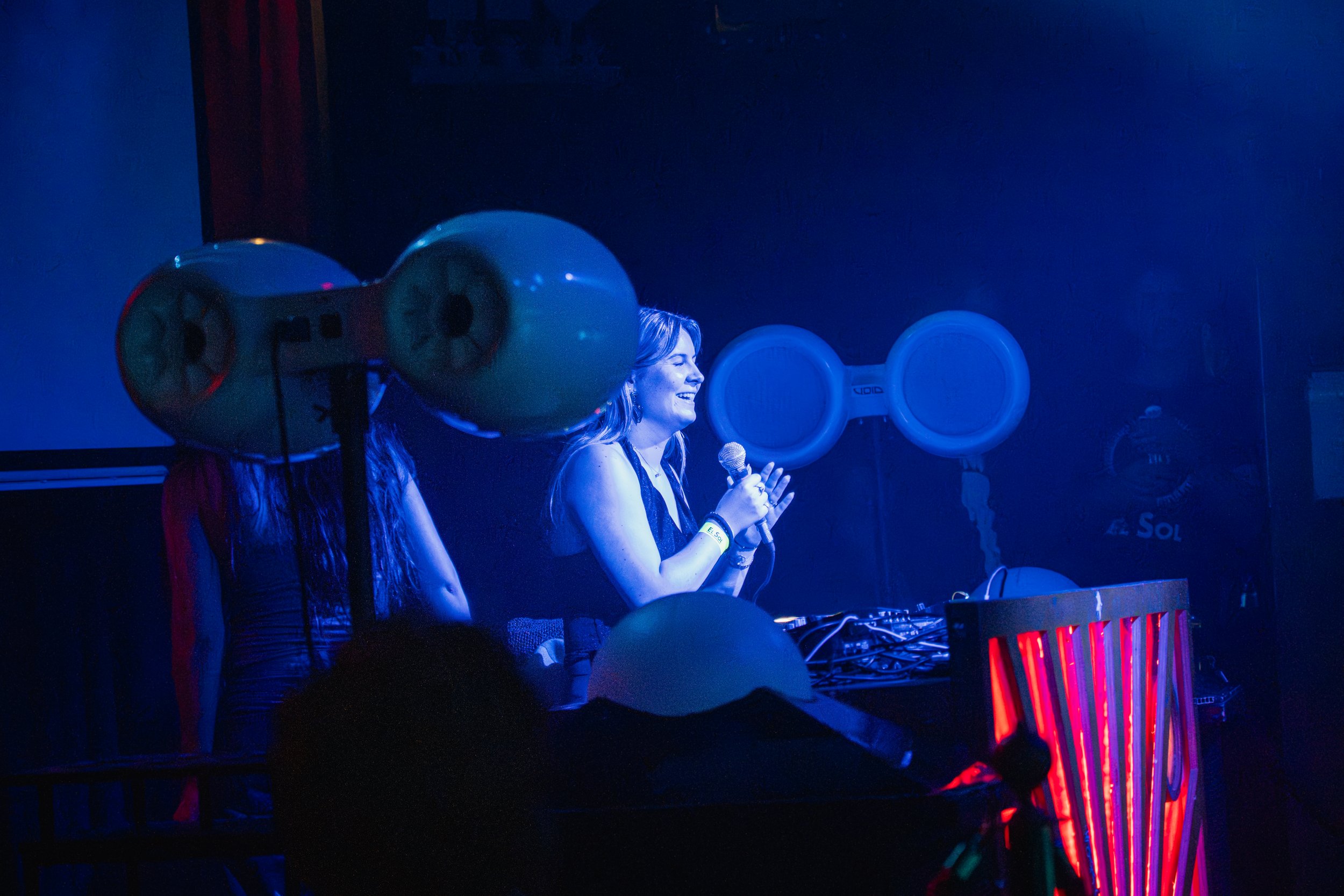 A woman smiling and speaking into a microphone behind DJ equipment at a nightclub or party, illuminated by blue and purple lights, with balloons and large speakers in the background.