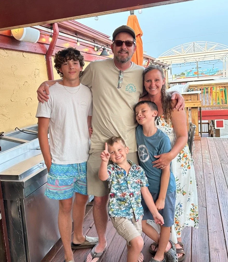 Family of six posing together at an outdoor location, with a picnic table and amusement park rides in the background.