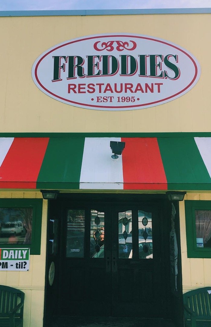 The exterior of Freddie's Restaurant with a yellow wall, a green, white, and red striped awning, and a black door. The sign indicates it was established in 1995.