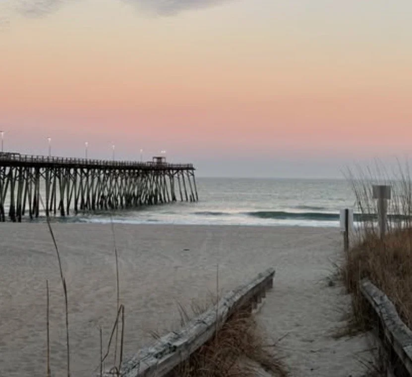 Beach scene with a wooden pier extending into the ocean during sunset, sandy pathway with grasses, and a clear sky with pink and orange hues.