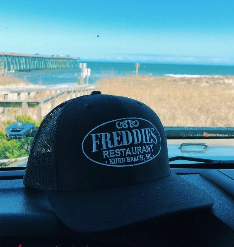A black trucker hat with white embroidered text that says 'Freddie's Restaurant, Kure Beach, NC' is on a vehicle dashboard. The background shows a coastal scene with a pier extending into the ocean, sandy dunes, and a blue sky.
