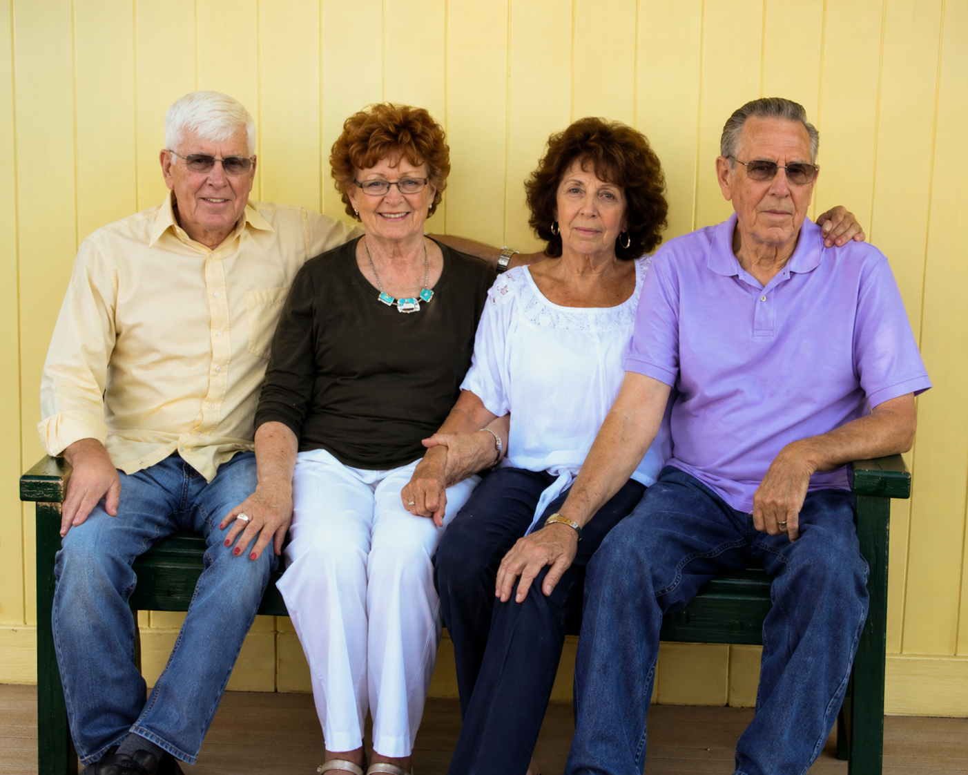 Four elderly people sitting on a green bench against a yellow wall, with two men and two women.