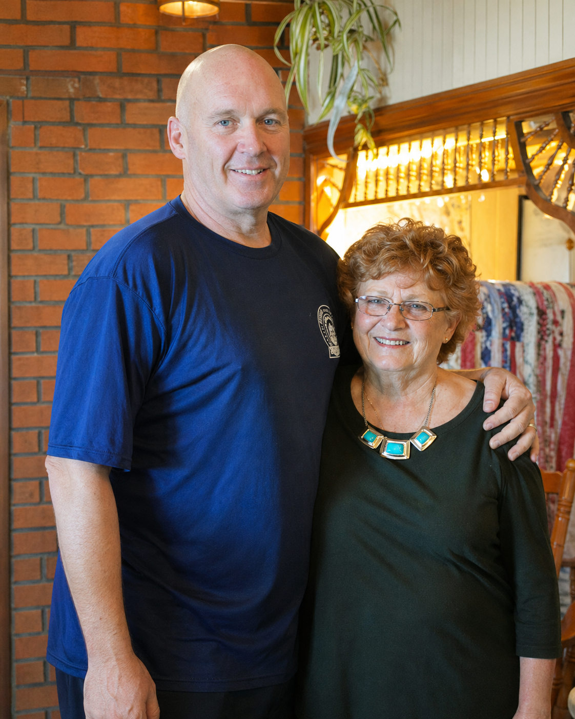 A man and a woman standing indoors, smiling and posing for a photo, with a brick wall, a plant, and a wooden railing in the background.