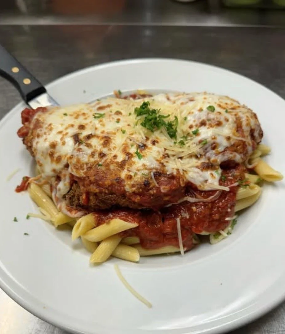 Plate of baked ziti pasta with marinara sauce, topped with melted cheese and garnished with chopped parsley, served with spaghetti underneath.