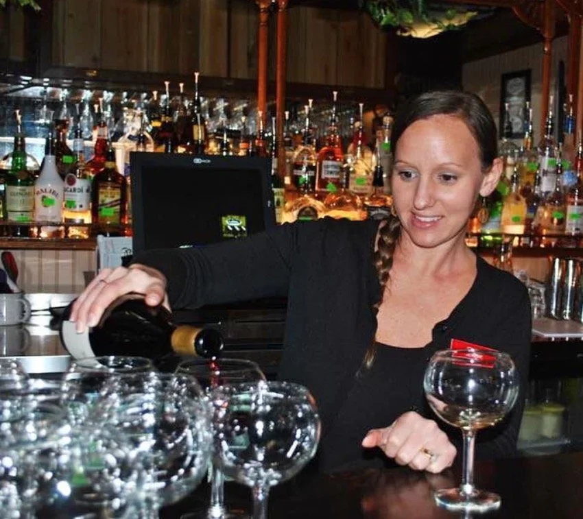A woman pouring red wine into a glass at a bar with various liquor bottles in the background.