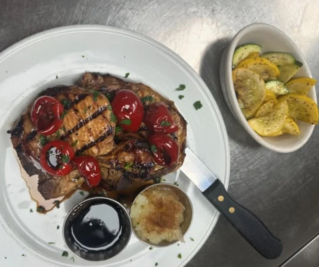 A plate with grilled steak topped with cherry tomatoes and herbs, served with two small bowls of sauces, and a side of mixed vegetables in a separate bowl.