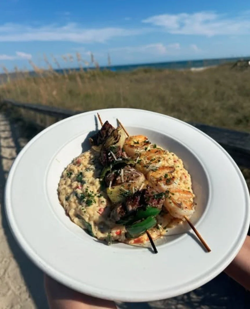 Plate of grilled shrimp skewers and rice with mixed vegetables, held outdoors near a grassy beach area with ocean view.