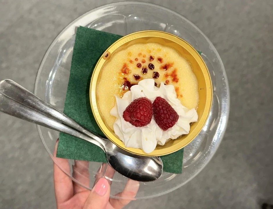 A small, round serving of custard topped with whipped cream and two raspberries, served in a yellow plastic container. The container is placed on a green napkin, on a clear glass plate, with a silver spoon beside it.