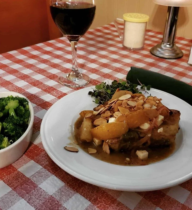 A glass of red wine, a plate of food with a dessert topping, a bowl of broccoli, and a candle on a checkered tablecloth.