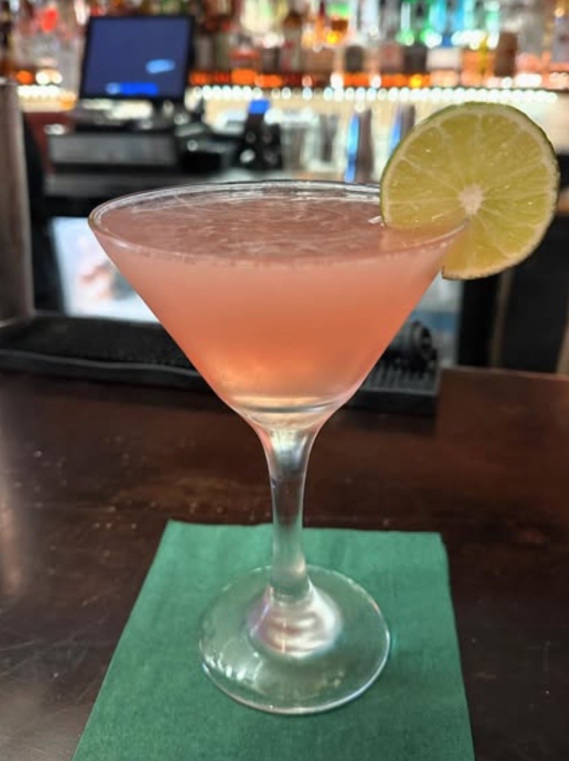 A pink cocktail garnished with a lemon wedge on a martini glass, placed on a green napkin at a bar.