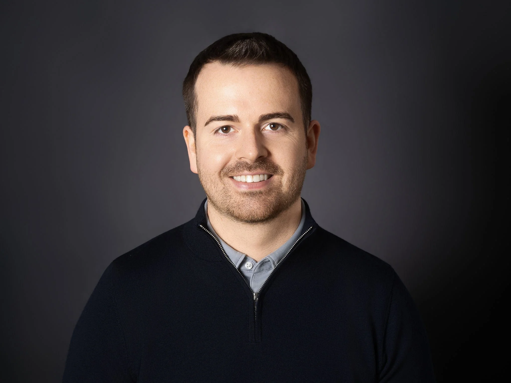Richard Pitcairn, Founder, Trusure, Portrait headshot of Richard, smiling face on at the camera, wearing a dark zip-up sweater with a light grey collared shirt underneath, against a dark background.