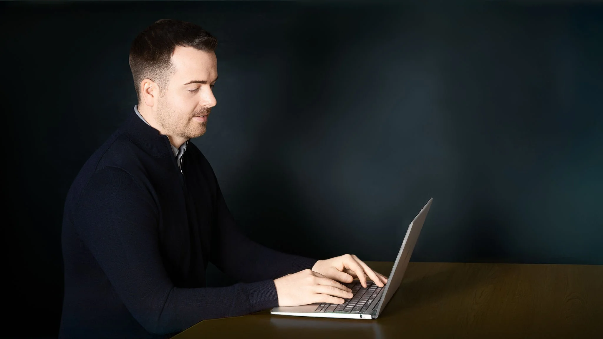Richard Pitcairn, Founder, Trusure , wearing a navy blue jumper, sitting at a wooden table, working at his laptop computer.