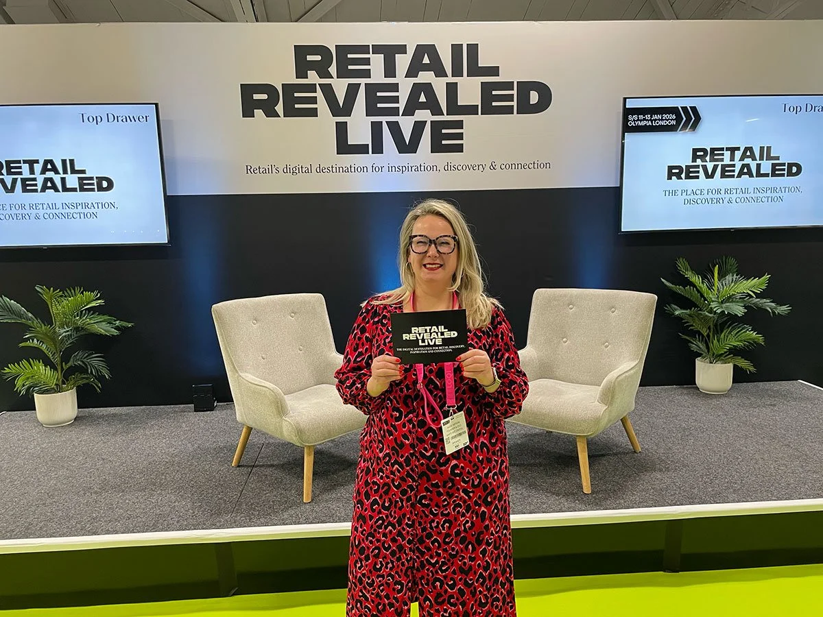 A woman in a red animal print dress holding a brochure stands in front of a stage with two beige armchairs and two potted plants. Behind her, a large sign reads 'Retail Revealed Live,' with screens displaying the same text.