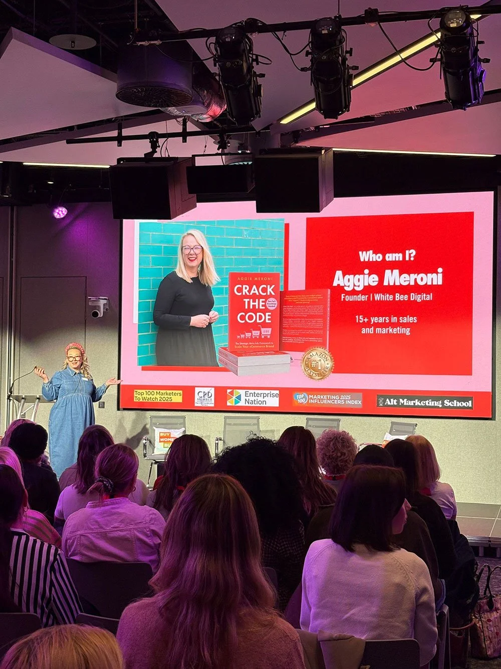 A woman is giving a presentation to an audience in a conference room. The large screen behind her displays a slide about Aggie Meroni, including her photo, a book titled "Crack The Code," and details about her background in marketing. The room is dimly lit with purple and pink lighting, and the audience is seated facing the stage.
