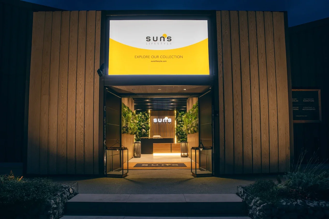 Store entrance with lit sign reading 'SUNS' inside and a large digital advertisement above the doorway, framed by wooden panel walls and greenery.