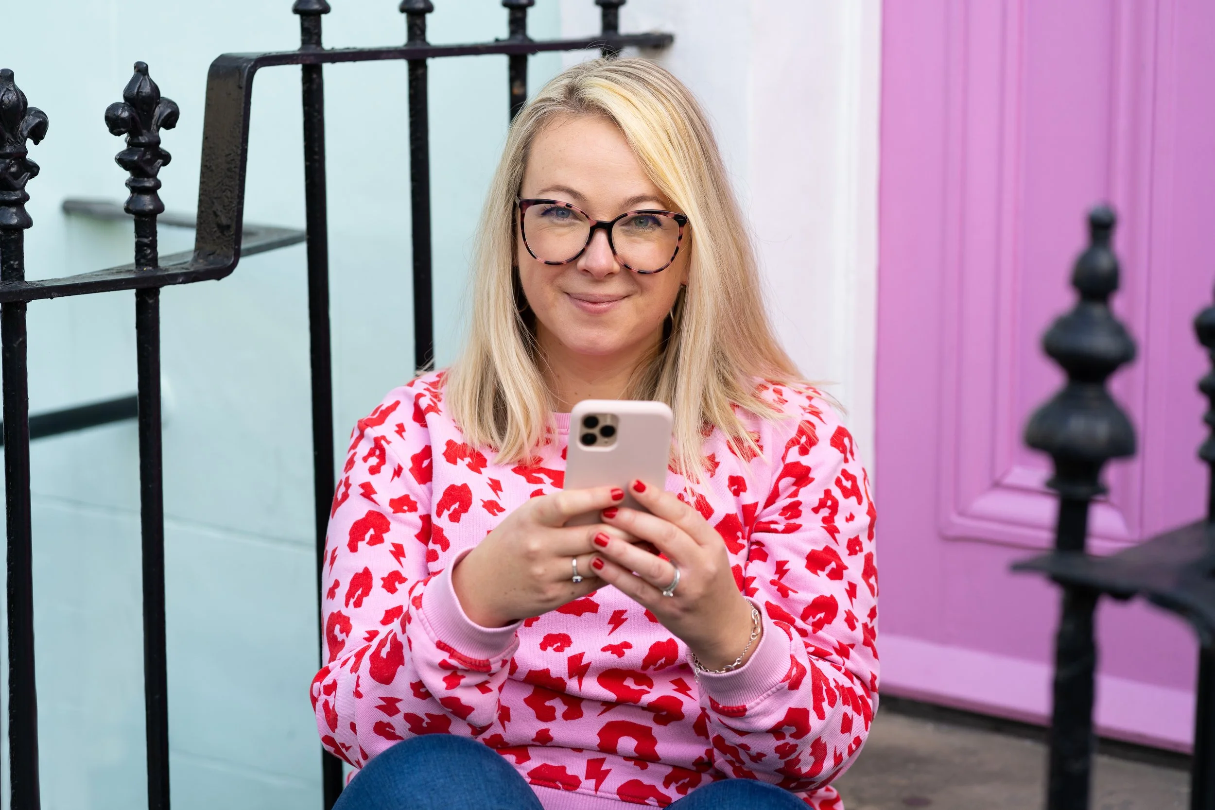 A woman with blonde hair and glasses looking at her phone, sitting outdoors near a pastel-colored building with a pink door, black iron railing, and white wall.