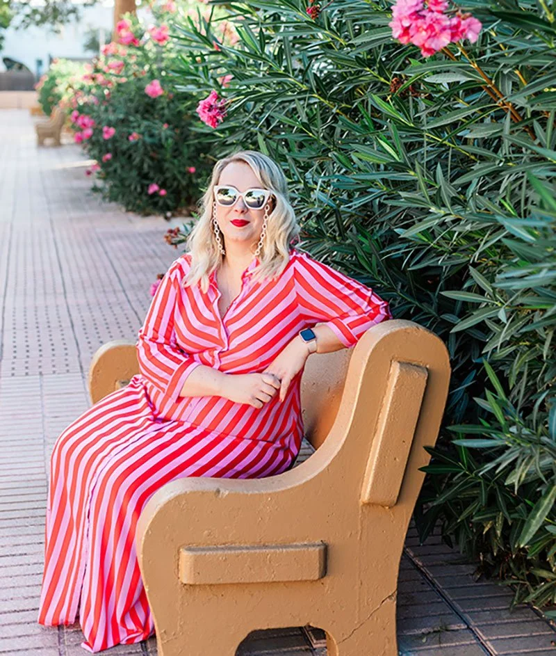 A woman with blonde hair, wearing sunglasses, red lipstick, earrings, a red and white striped dress, and a watch, sits on a bench next to green bushes with pink flowers.