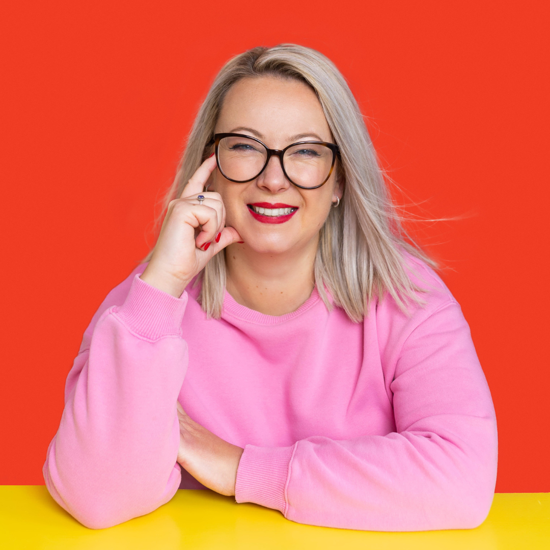 A woman with blonde hair, glasses, and red lipstick, wearing a pink sweatshirt, smiling and winking, sitting against a red background with a yellow surface in front.