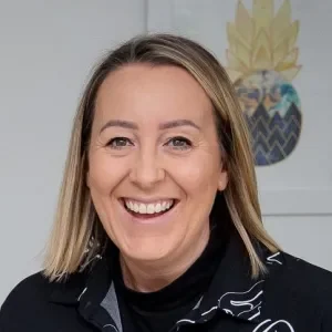 A woman smiling with shoulder-length blonde hair, wearing a dark patterned top, standing in front of a light-colored wall with a decorative pineapple artwork.