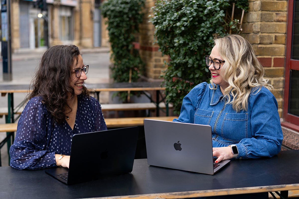 Two women sitting at a table outdoors, smiling and engaging in conversation, with laptops in front of them.