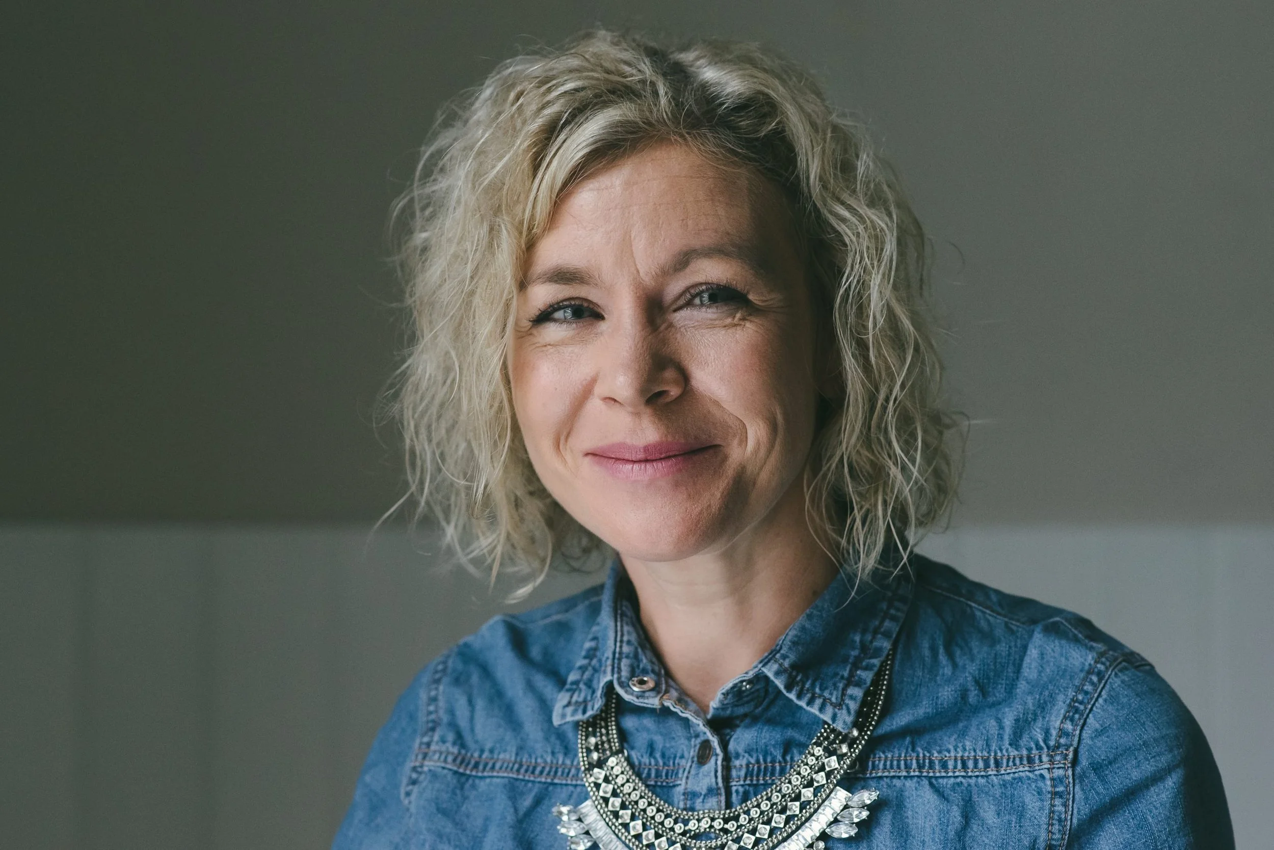 A middle-aged woman with curly blonde hair, wearing a denim shirt and a statement necklace, smiling softly with a neutral gray background.