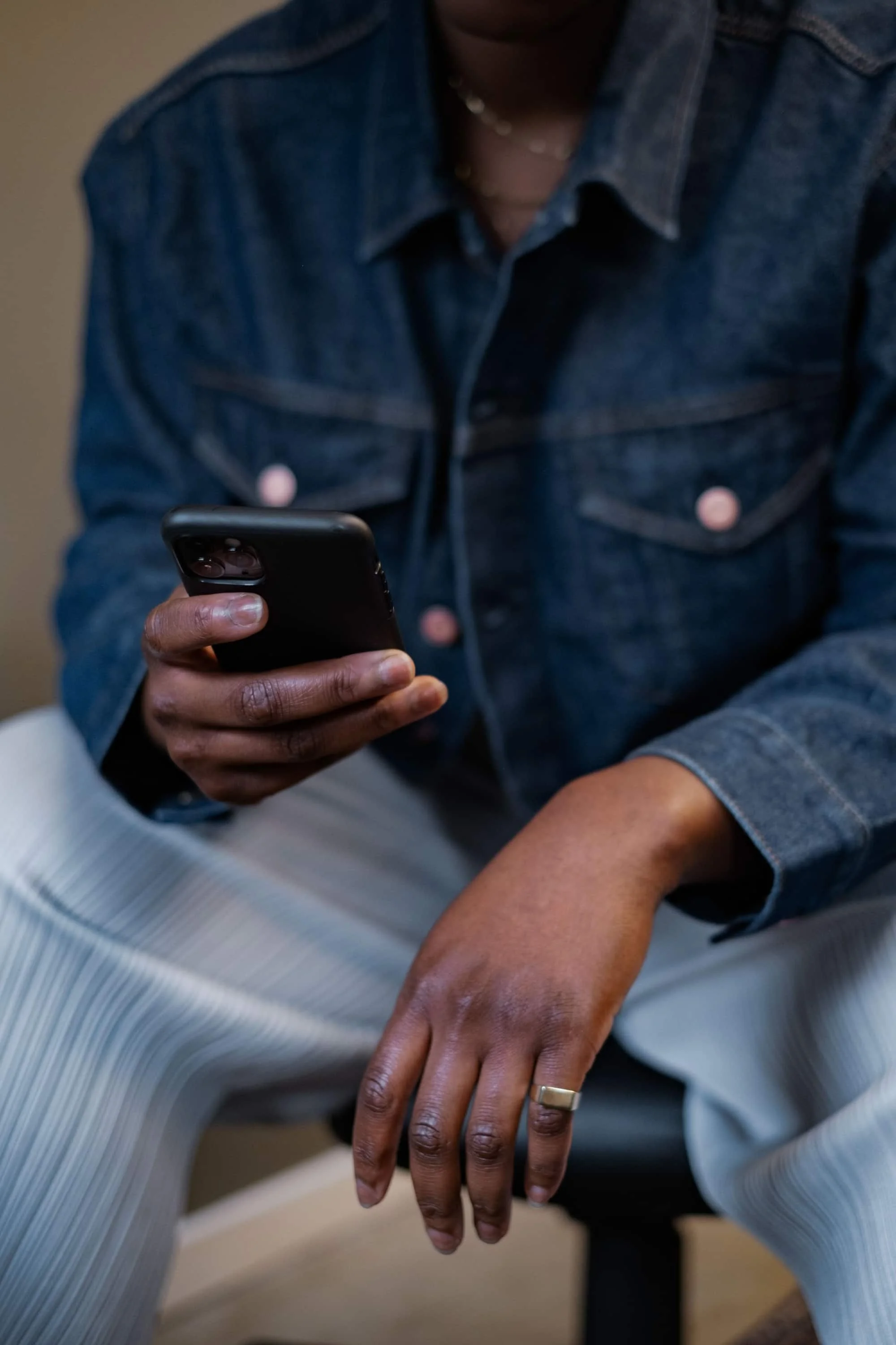 Close-up van een persoon die een smartphone vasthoudt, met een hand met een ring op de vinger die rust op de knie van een andere persoon met een vrij donkere huid.