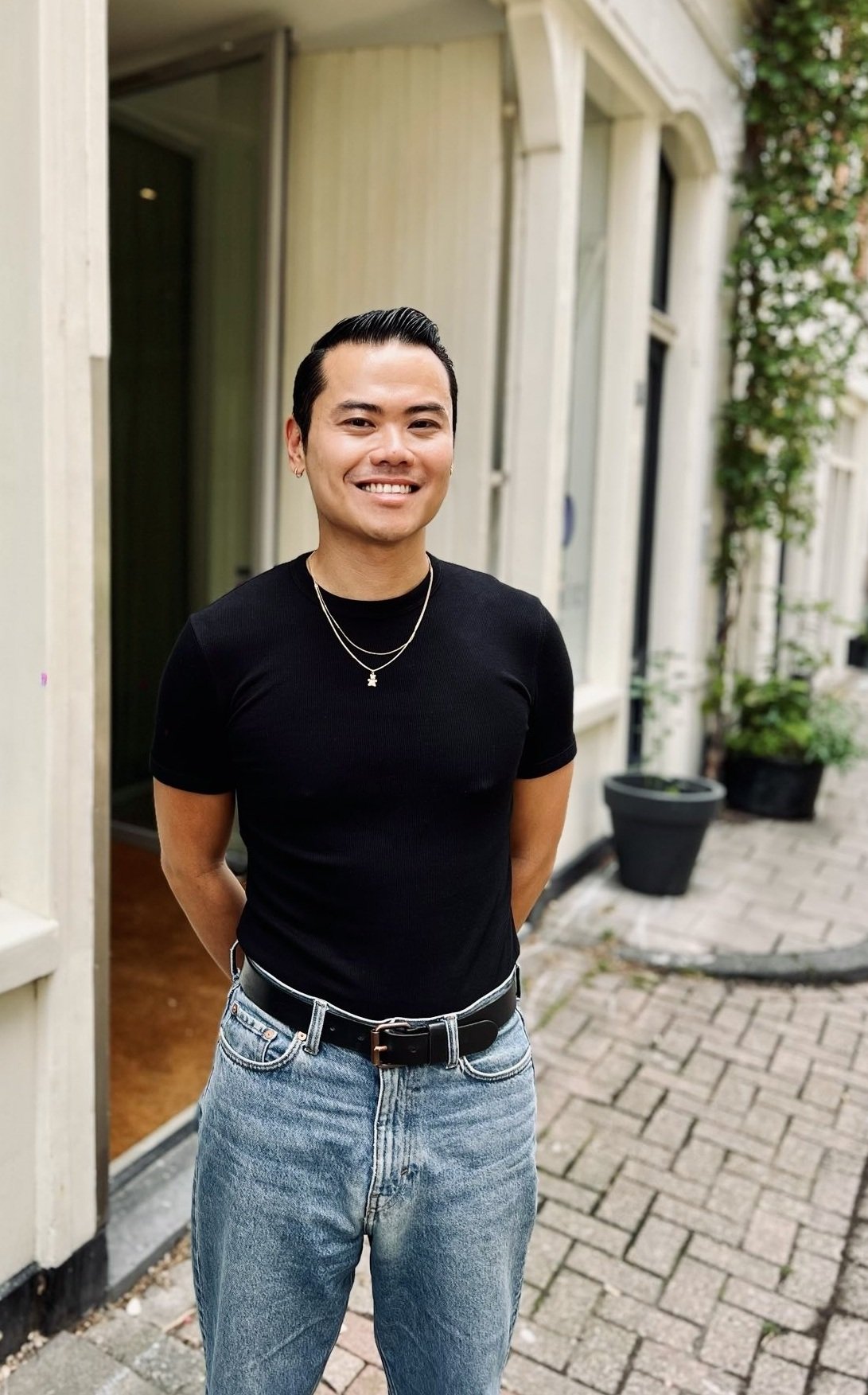 Een lachende jonge man met zwart haar, in een zwart shirt en blauwe jeans, staat buiten op een straat met planten en behuizingen op de achtergrond.