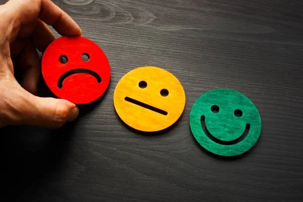 Hand die een rood, een geel en een groen smiley op een donkere tafel vasthoudt, die de emoties verdrietig, neutraal en blij uitdrukken.