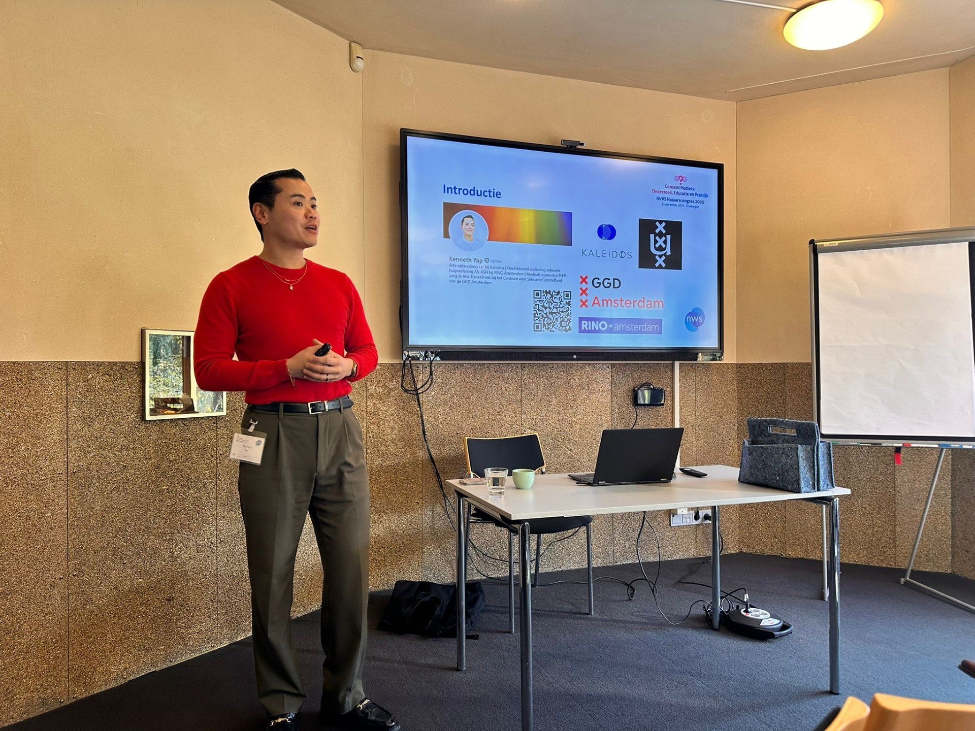 Man in rode trui geeft presentatie in vergaderruimte met scherm en witteboard.