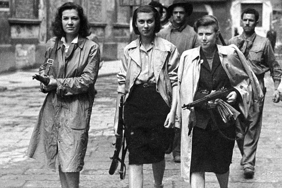 Black and white photograph of five women walking down a cobblestone street, carrying rifles, dressed in casual clothing, with somber expressions.