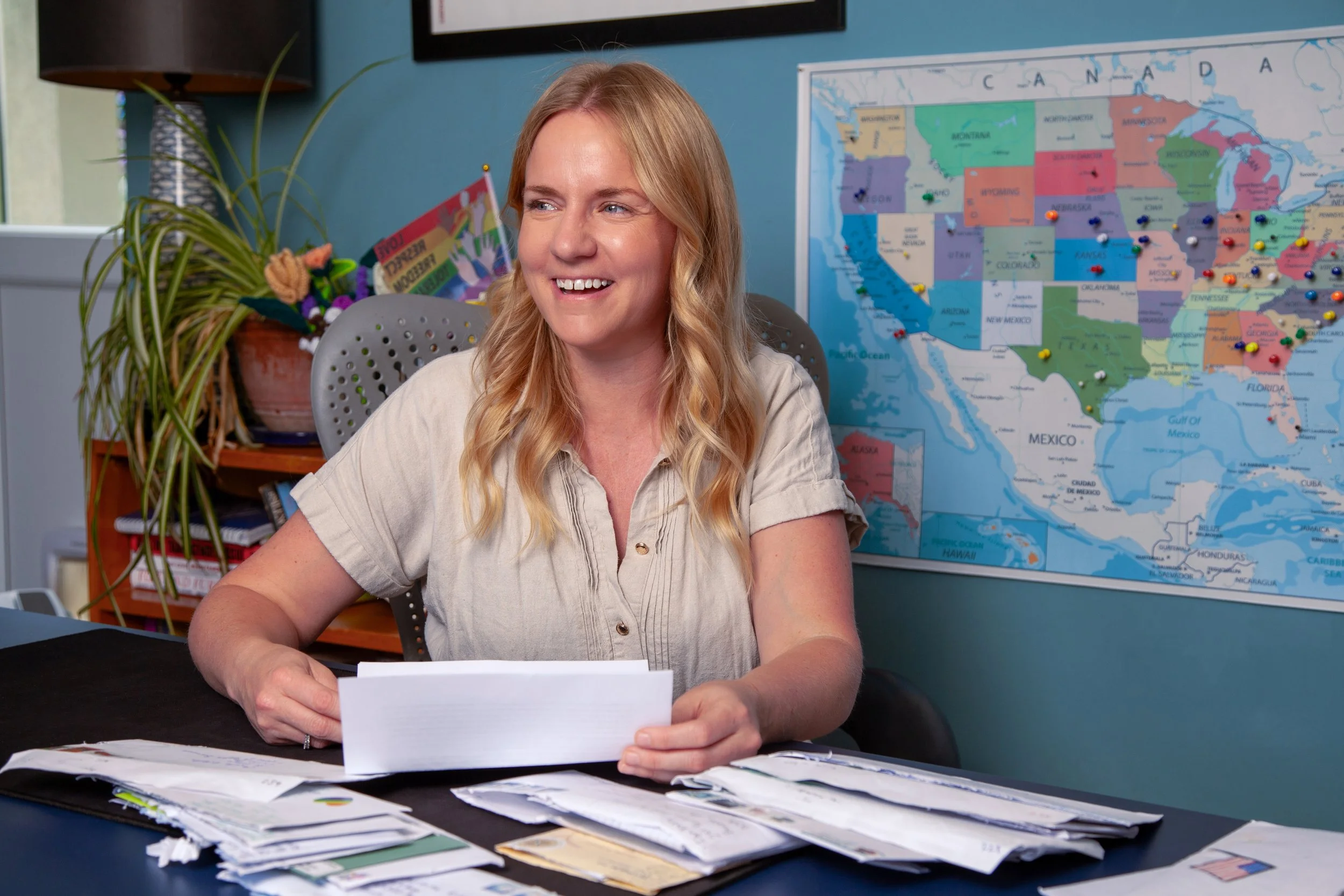 A woman with blonde, wavy hair smiling as she looks to her left while sitting at a desk covered with papers and notes, with a large map of the United States on the wall behind her and a potted plant nearby.