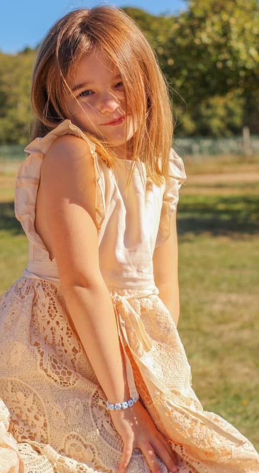 A young girl with long red hair smiling outdoors in a park on a sunny day, wearing a cream-colored dress with lace details.