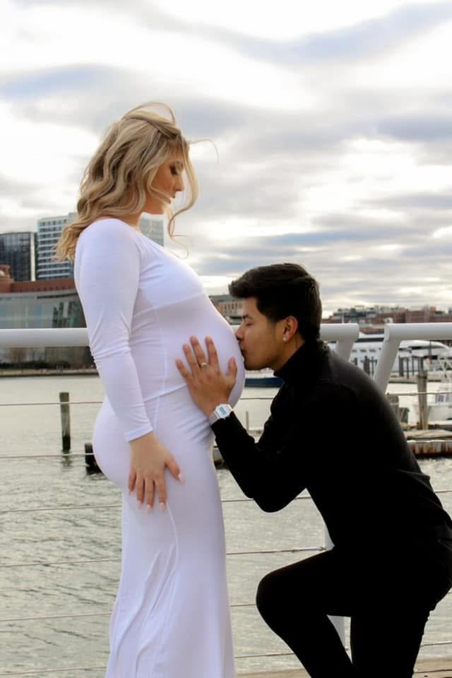 A man kneeling and kissing a pregnant woman’s stomach outdoors near a body of water with city buildings in the background.