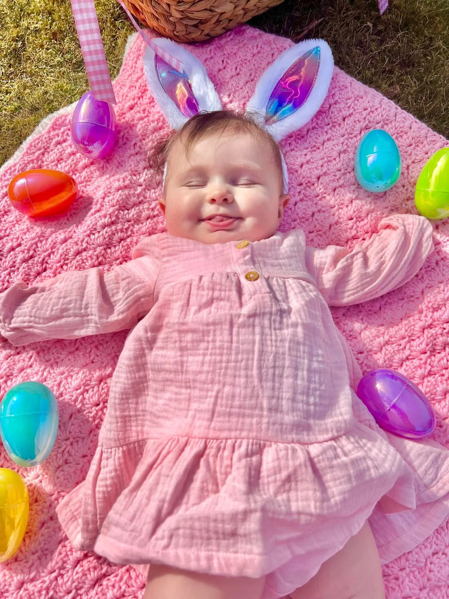 A smiling baby girl with bunny ears lying on a pink blanket outdoors, surrounded by colorful plastic Easter eggs.
