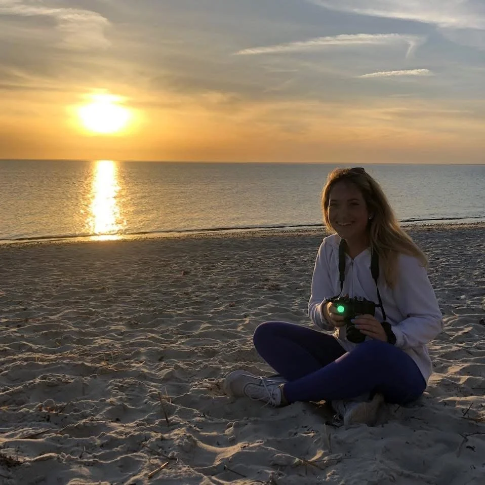 A woman sitting on the sandy beach at sunset, holding a camera, with the ocean and colorful sky in the background.