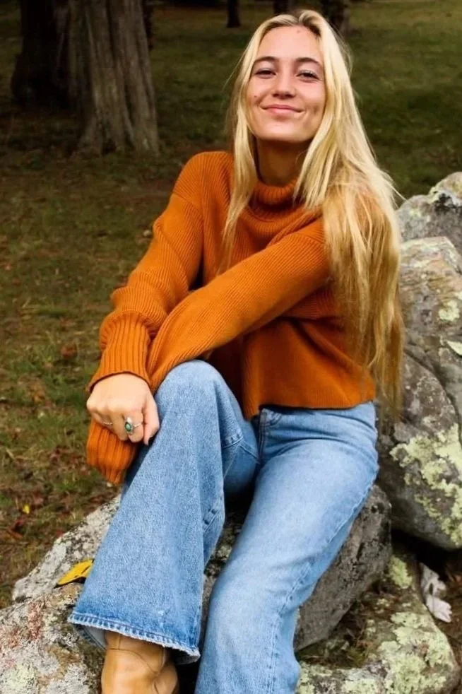 A young woman with long blonde hair smiling, sitting on a rock outdoors with trees and grass in the background, wearing an orange sweater, blue jeans, and tan boots.