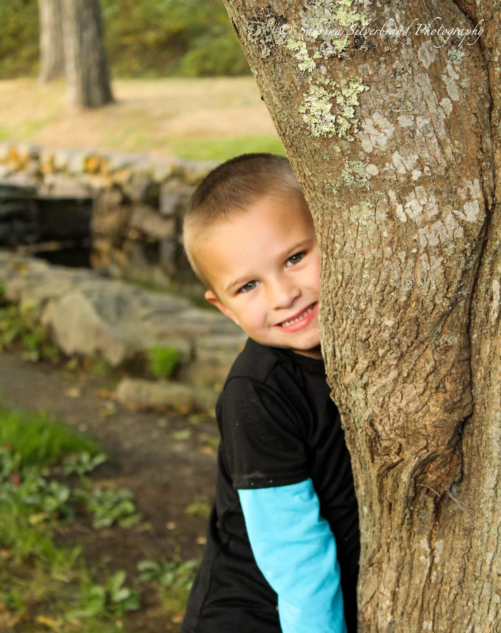A young boy with short hair and a big smile hiding behind a tree. The background shows a blurred outdoor park scene with greenery and a stone wall.