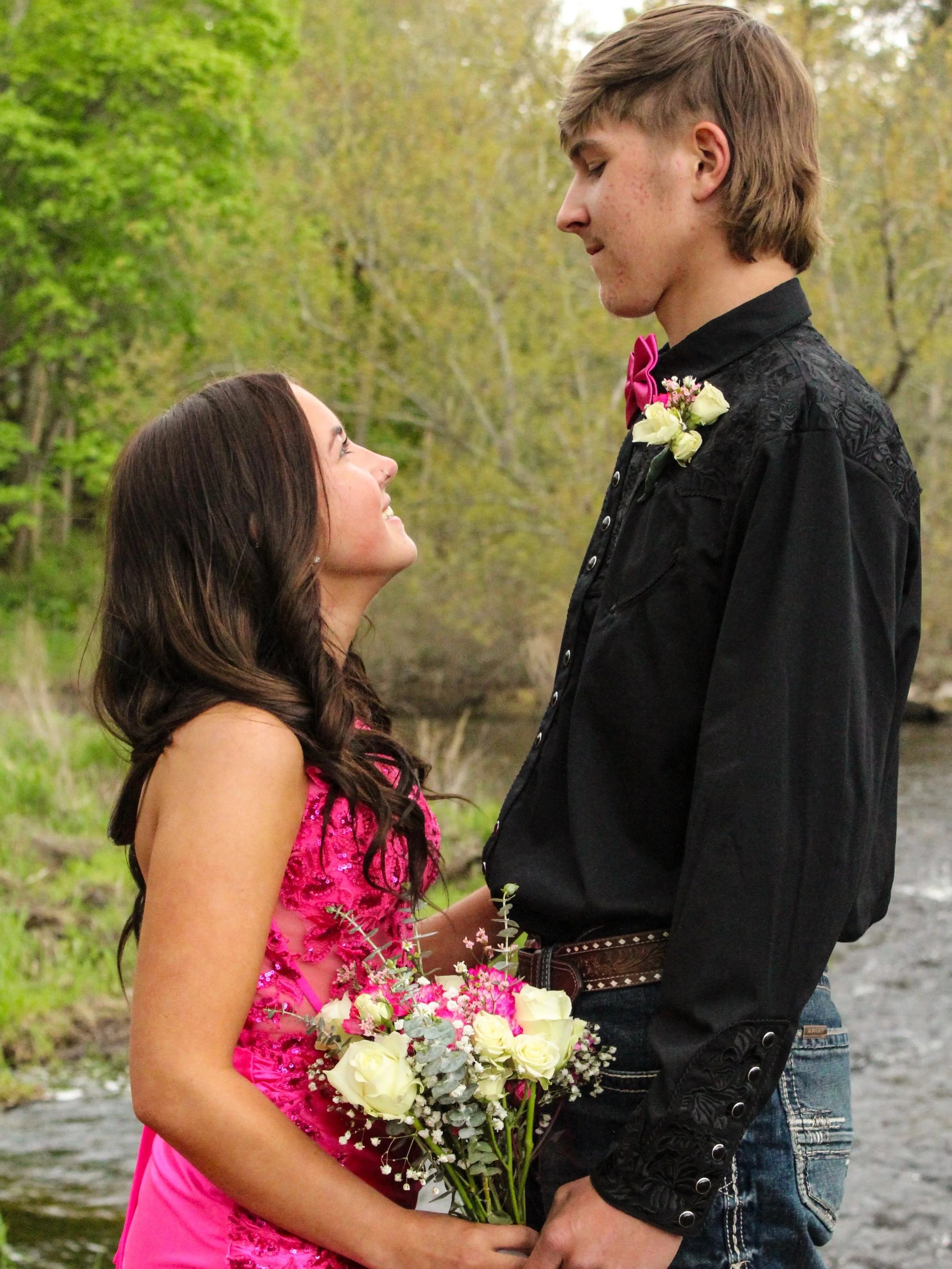 A young woman in a bright pink dress holding a bouquet of white and pink flowers, gazing up at a young man dressed in a black shirt with embroidered patterns, a pink bow tie, and a small boutonniere, outdoors with trees and a stream in the background.