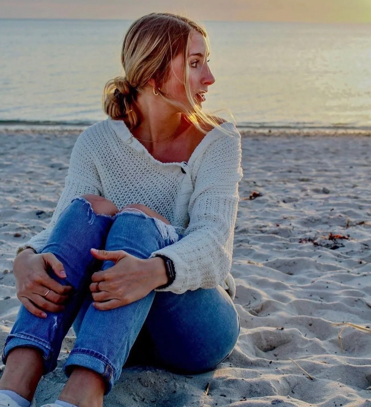 A woman sitting on the sand at the beach, hugging her knees, looking to the side during sunset.