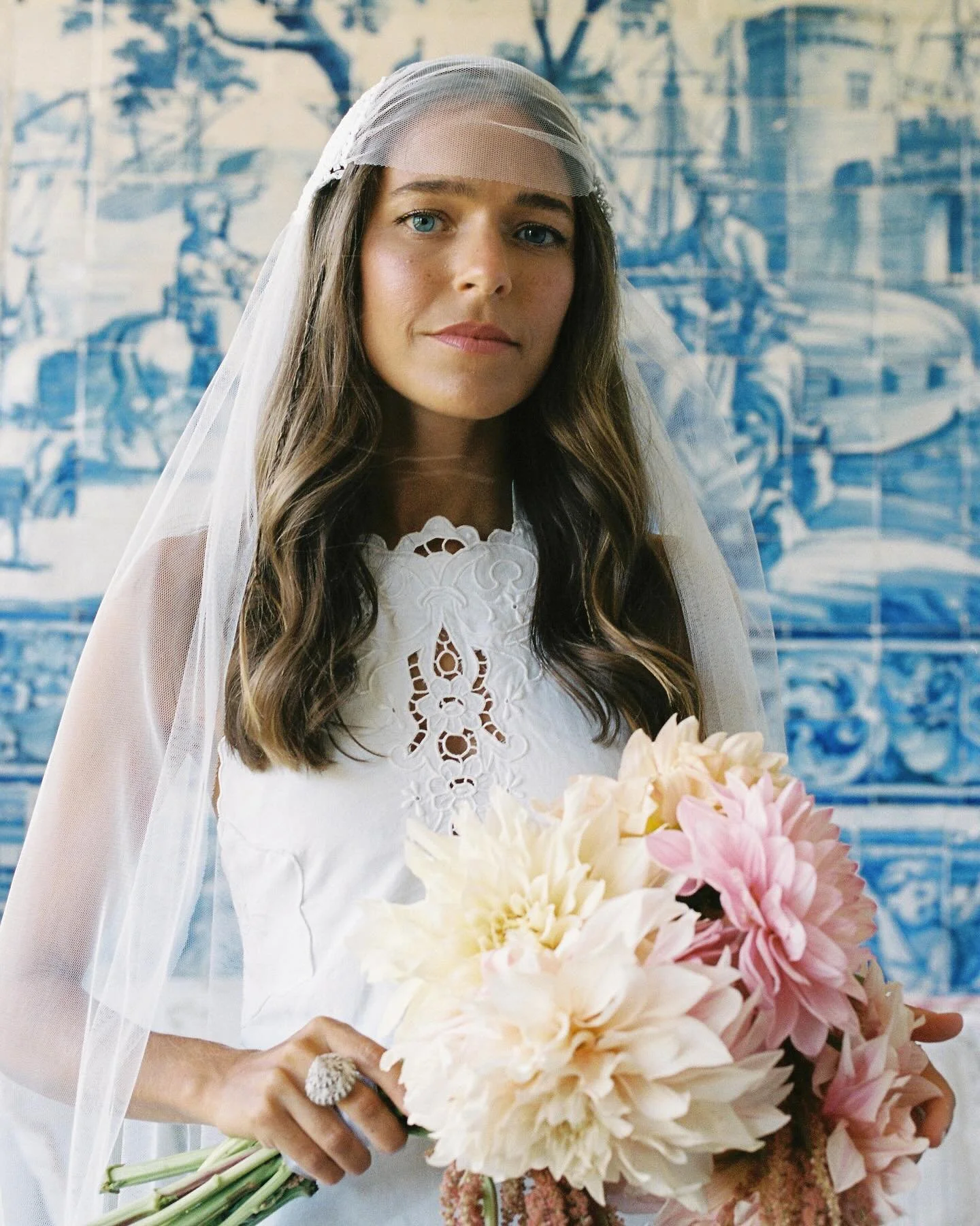 A woman dressed in a wedding gown and veil holding a bouquet of pink and white flowers, standing in front of a blue mural.