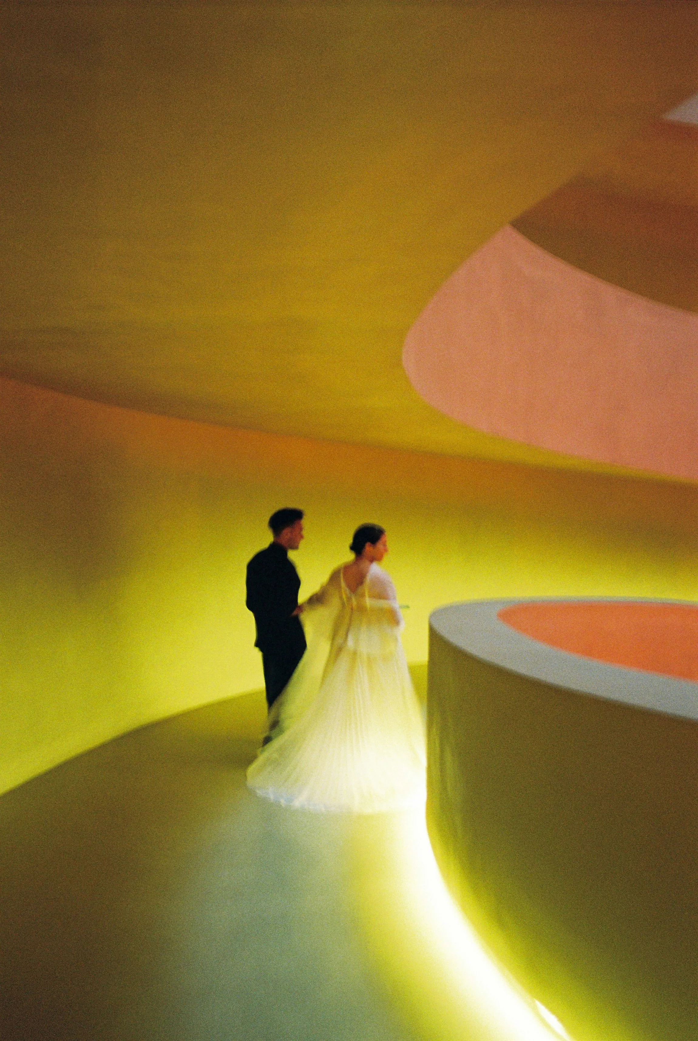 Two people, a man and a woman, standing in front of a vibrant yellow and pink curved abstract background. The woman appears to be wearing a flowing dress, and the man is dressed in dark clothing.