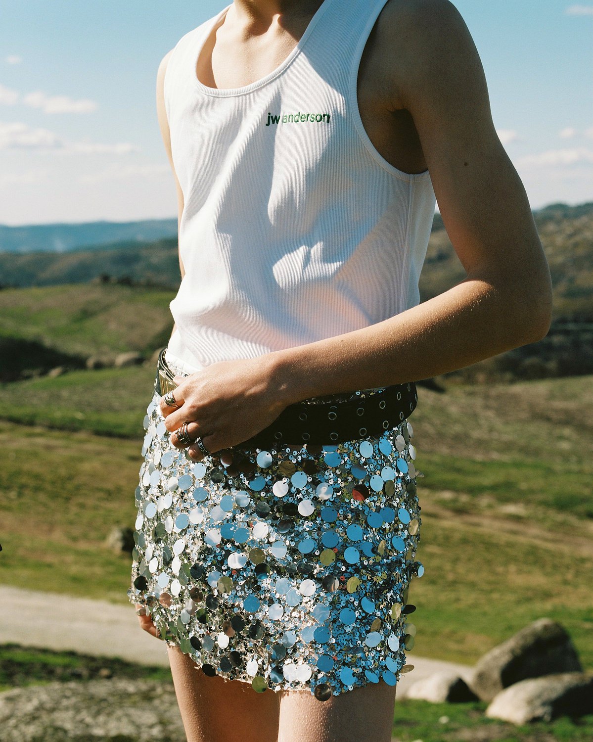 Close-up of a woman wearing a white tank top with green text and a shiny sequin skirt outdoors with a landscape background.