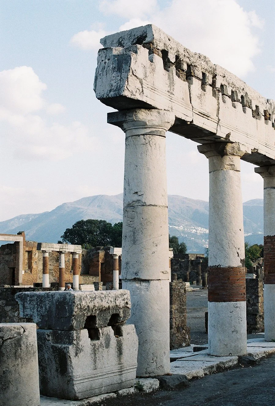 Ancient ruins of stone columns and structures, with mountains in the background and a partly cloudy sky.