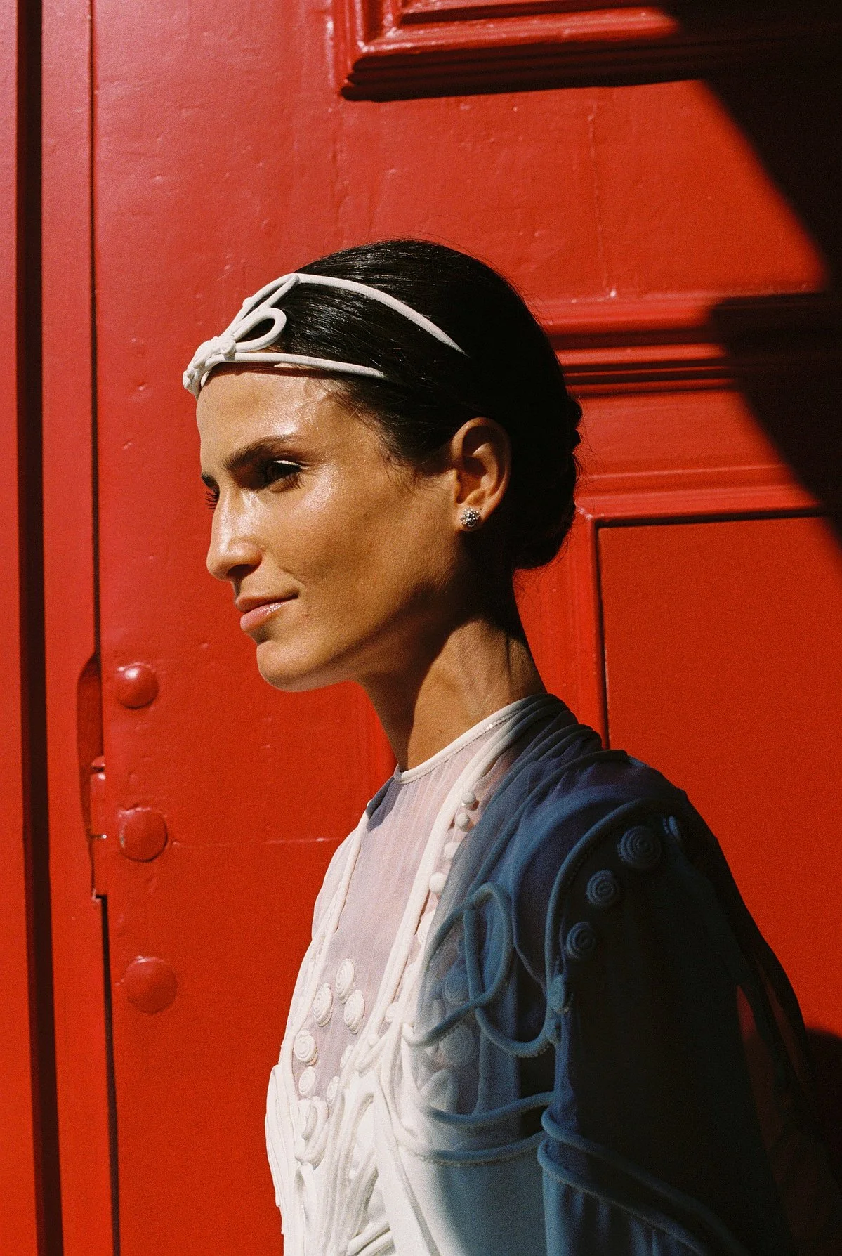 A woman with black hair styled in a sleek bun, wearing a white headband and earrings, standing against a red background. She's dressed in a white top and a blue jacket with circular detailing on the shoulder.