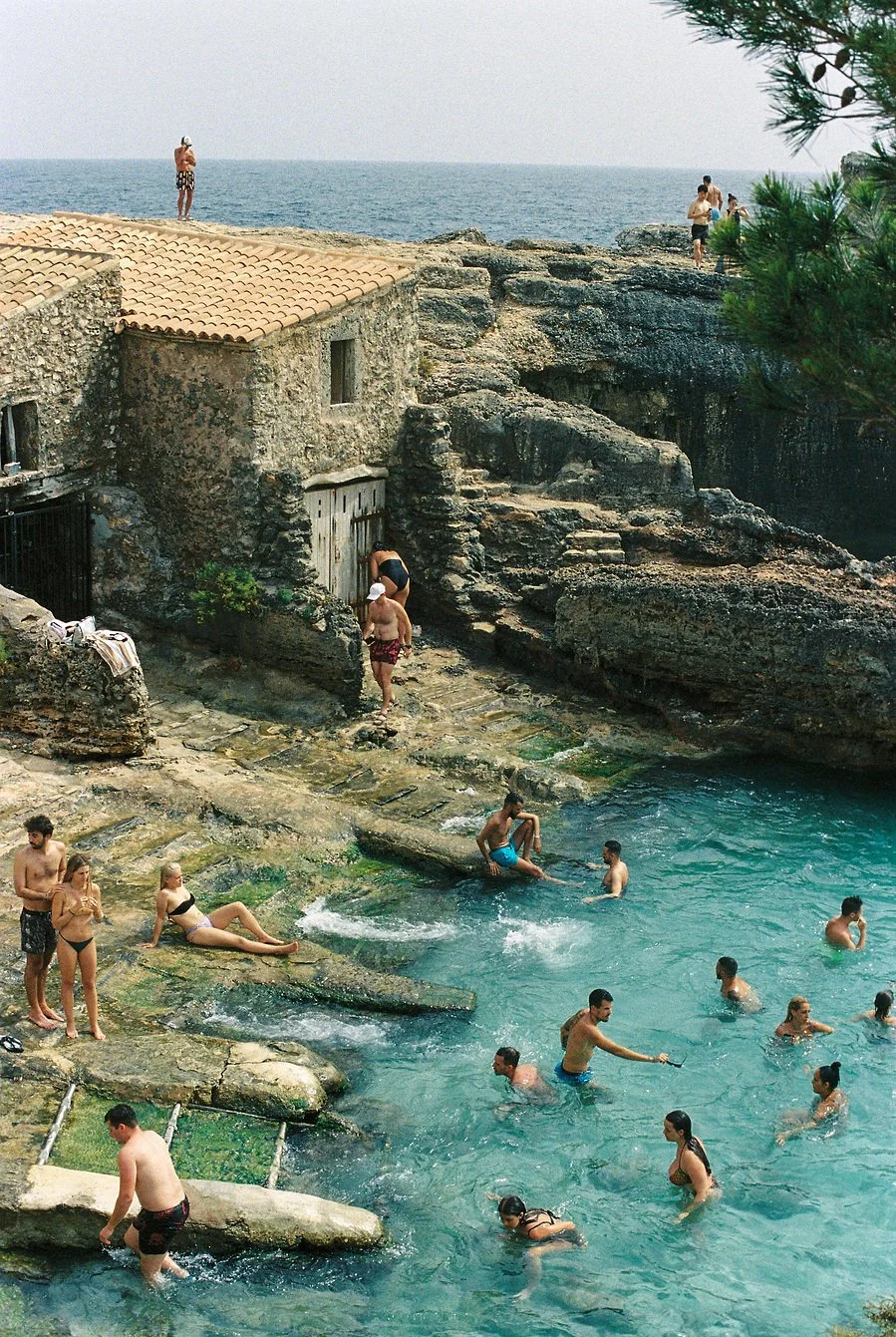 People enjoying a rocky seaside swimming area with a small stone house and ocean in the background.