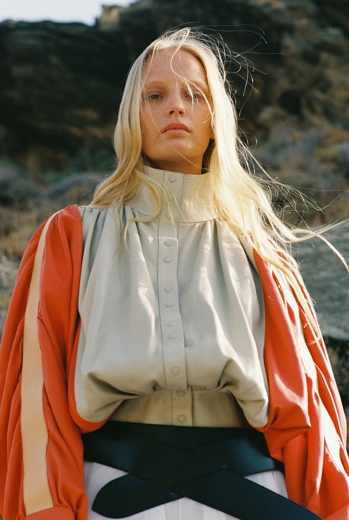 A woman with long blonde hair stands outdoors against a rocky background, wearing a high-neck beige blouse and a bright orange jacket draped over her shoulders.